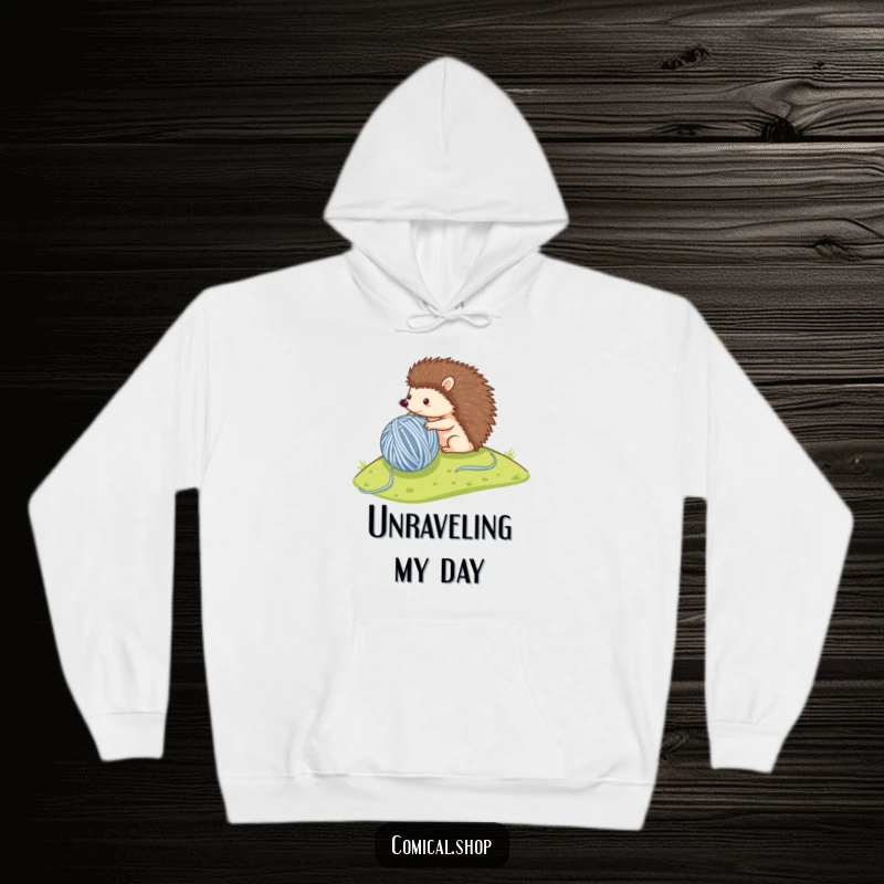 Funny hoodie depicting a small hedgehog determinedly rolling a massive ball of yarn down a hill, exuding cozy charm.
